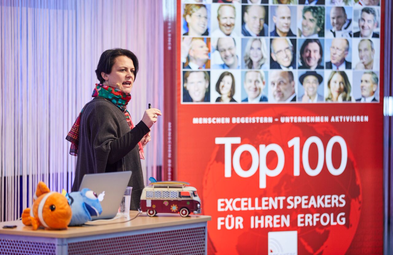 Stefanie Aufleger bei einem Vortrag neben dem Aufsteller Top 100 Excellent Speakers.
